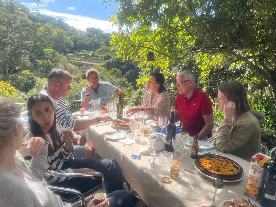Guests seated along a long table in the garden, mid-meal, with the wooded valley behind them.