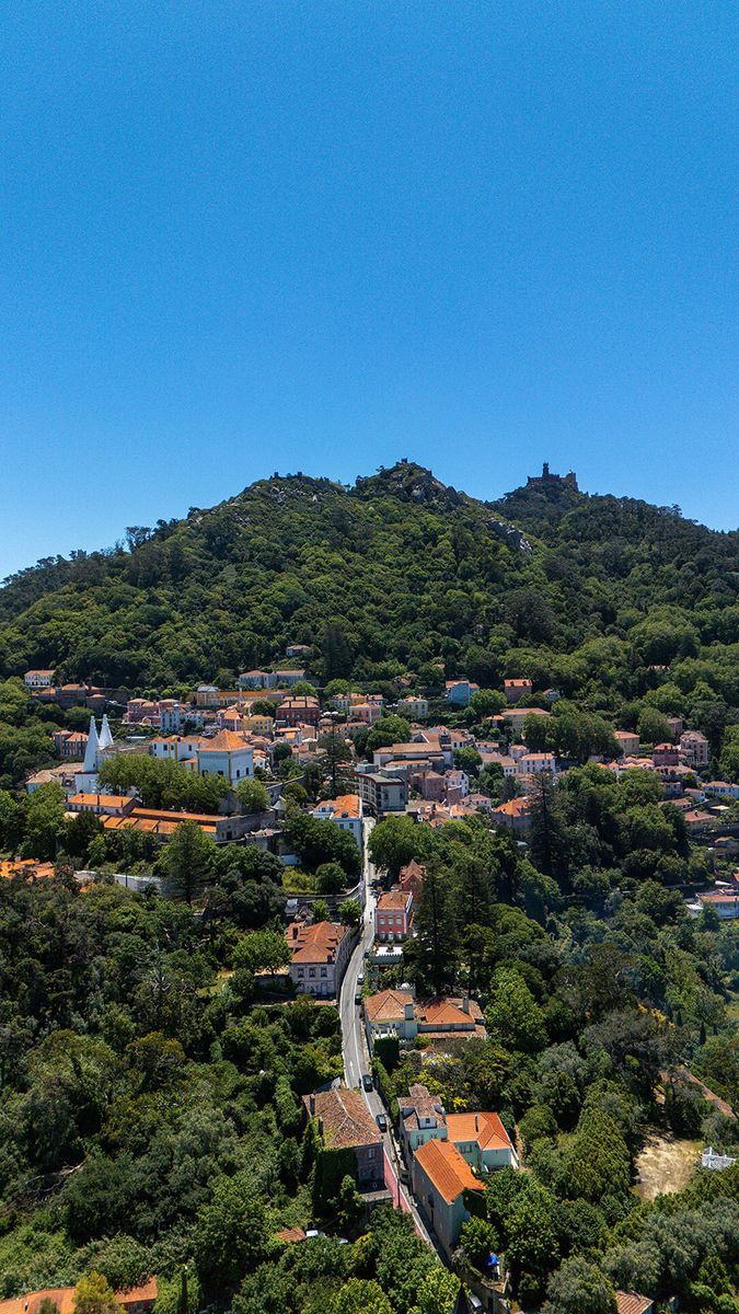 Sintra Old Town from the air: pastel houses and terracotta roofs packed under the wooded ridge, with the Moorish castle running along the top of it.